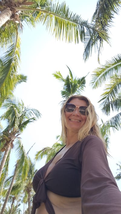 A woman wearing sunglasses and a dark brown crop top stands looking up at the camera, smiling. Palm trees surround her, framing the sky. The location is not discernible from the image to be in the Jiyang District of China.