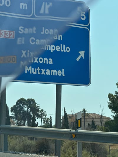 This media file shows a road sign in Spanish, likely in or near Mutxamel, Spain. The sign is blue with white text and an arrow pointing left. Visible text includes "Sant Joan," "El Campello," "Xixona," and "Mutxamel." A number "5" is also visible on a separate, adjacent sign. Below "Sant Joan" and "El Campello," the number "332" is displayed in a red rectangle. Further down, "Mutxamel" is preceded by "N." The sign appears to be mounted on a metal post, with a guardrail to the right.
In the background, there is lush green vegetation, including trees and palm trees. A building with a tiled roof is visible behind some of the trees. The sky is clear and appears to be a bright, possibly sunny day, suggesting daytime. The scene is viewed from a vehicle, as indicated by the blurriness and the angle of the shot, implying the subjects are traveling. The overall impression is of a roadside scene in a Mediterranean climate.