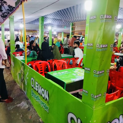 This media file captures a vibrant, temporary dining or refreshment area, likely part of an event or market in Charfasson, Bangladesh. The setting is dominated by a bright green color scheme, featuring green pillars and table coverings prominently displaying the brand "CLEMON." The canopy overhead is a white and blue checkered pattern, supported by yellow poles, suggesting a tent-like structure. Artificial lighting from hanging lamps indicates it is likely evening or night.

Numerous people, including adults and children, are present. They are casually dressed, with several women wearing head coverings. Individuals are seen seated at tables on red plastic chairs, engaging in conversation, eating, or observing their surroundings. The overall atmosphere appears lively and relaxed, with people enjoying the social interaction.

Key objects include the red plastic chairs and green tables, many of which are covered with branded tablecloths. The green pillars and table skirts are adorned with the "CLEMON" logo in white, often accompanied by Bengali text that primarily reads "ক্লিয়ারলি লেমন" (Kliyārli Lemon), meaning "Clearly Lemon." Some variations like "লি লেমন" (Li Lemon) are also visible. A shelf stocked with bottles is seen in the background, likely part of a service counter. On the ground, which appears to be dirt or concrete, a piece of plastic litter is visible near the base of the branded enclosure.

The scene suggests a casual gathering place, possibly a food stall at a local fair or a promotional setup for the "CLEMON" beverage brand, offering a space for refreshment and socializing.