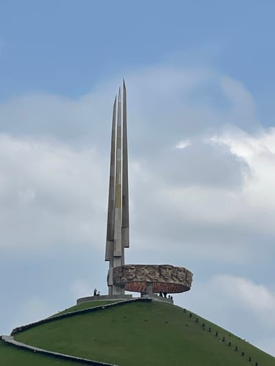 This is a vertical, medium shot of the Victory Monument in the Ozyaritska-Slabadski rural council, Belarus. The monument is a tall, sharp, needle-like structure made of concrete and metal, with three distinct triangular levels converging at a point. To the right of the needle, there's a large, circular sculpture with intricate carvings and what appears to be stylized figures. This circular sculpture is mounted on a concrete base. Beyond the monument, a green, sloped hill rises, covered in grass and with a dark line running along its edge, possibly indicating a path or structure. There are several small, dark figures scattered on the green hill and near the base of the circular sculpture, which are likely people. The sky is a pale blue with scattered white clouds, suggesting it is daytime. The overall impression is one of grandeur and memorial.