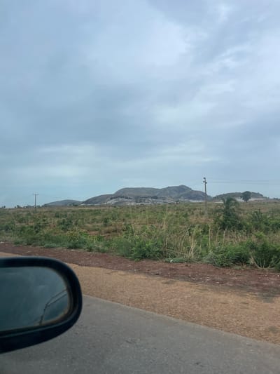 The image shows a landscape in Kujama, Nigeria, viewed from what appears to be the interior of a vehicle. A cloudy sky dominates the left portion of the image. In the center, there's a view of distant, grassy terrain leading to a rocky hill or small mountain. There is a utility pole with power lines. To the right, a narrow strip of roadside dirt and vegetation is adjacent to a paved road surface. In the bottom right corner, a side mirror of the vehicle is visible.