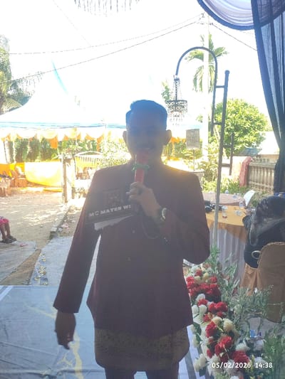A man is standing in front of a flower arrangement and holding a microphone. He wears a dark red jacket and a patterned skirt. The background shows an outdoor event with a white tent and some decorative lighting. A table with a white tablecloth and water bottles is visible behind the man. The date 05/02/2026 and the time 14:38 are shown in the lower right corner.