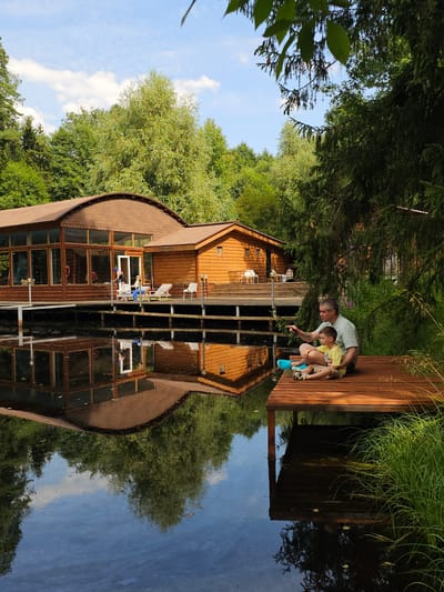 A man and a boy are sitting on a wooden pier on a body of water, with a wooden building with a curved roof in the background. The man, wearing a light-colored t-shirt and pants, is pointing his left index finger towards the water. The boy, wearing a yellow t-shirt and shorts, is sitting next to him, looking in the same direction. They are both seated on the pier with their legs crossed. A blue flotation device is placed near them on the pier. The water reflects the building and trees. The building appears to be a lodge or resort, with large windows and wooden siding. The surrounding environment is lush with green trees and foliage. This scene is located in the village of Ulitkino, Russia.