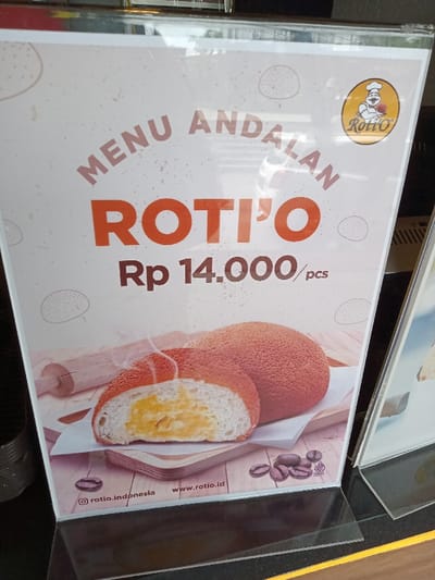 An eye-level shot captures a display stand featuring an advertisement for "Roti'O" buns, likely placed in a food establishment in Kecamatan Panji, Indonesia. The stand is made of clear acrylic, causing slight reflections on the surface.
The poster prominently displays two round, crusty buns with a distinctive texture. One bun is cut in half, revealing a rich, creamy yellow filling and subtle steam rising, suggesting warmth and freshness. In the background of the illustration, a wooden rolling pin is visible, hinting at the bakery process. Towards the bottom right, a scattering of coffee beans is depicted, potentially indicating a coffee-flavored crust or a suggested pairing.
Key text elements are clearly visible:
- "MENU ANDALAN" (meaning "Featured Menu" or "Signature Menu" in Indonesian) is written in an arched fashion at the top.
- The brand name "ROTI'O" is prominently displayed in large orange letters below.
- The price "Rp 14.000 / pcs" (14,000 Indonesian Rupiah per piece) is shown beneath the brand name.
- In the top right corner, a circular logo features a cheerful chef character holding a bun, with the "Roti'O" name.
- At the bottom left, the Instagram handle "@rotio.indonesia" and the website "www.rotio.id" are listed.
- A "HALAL" certification logo in Arabic script is also visible near the coffee beans, confirming the product's adherence to Islamic dietary laws.
The overall impression is that of a professional and inviting advertisement for a popular bakery item.
