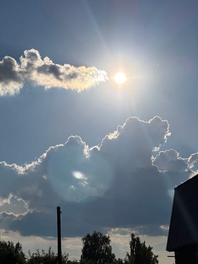 The image depicts a sky filled with dramatic clouds illuminated by the sun. In the upper portion of the frame, the sun is a bright orb, partially obscured by a thin, wispy cloud band that shows iridescence with hints of yellow, orange, and blue. Below this, larger, darker clouds form massive formations with bright, illuminated edges, suggesting they are catching the sunlight from behind. In the lower part of the image, silhouetted against the sky are trees and the dark roof of a building. A single, dark wooden pole stands vertically in the mid-ground. The overall scene is one of a dynamic sky, likely captured in the town of Krivandino, Russia, given the context. There are no people or animals visible in the frame.