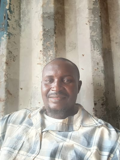 A portrait-style photograph captures an adult Black male, likely in Iddah, Nigeria, smiling faintly and looking directly at the camera. He has short dark hair and a short, dark beard or goatee. He wears a light-colored plaid button-up shirt, with checks in shades of white, blue, and beige, over a white undershirt.
The background is a weathered, corrugated metal wall, possibly part of a shipping container or an industrial building. The wall shows extensive peeling paint in light grey, white, and faint blue tones, revealing rust and general discoloration. Faint, hand-drawn or scratched text and symbols are visible on the wall, including what appears to be "Dilly" and "2019" with a triangular design to the upper left of the man's head.
A digital timestamp "2026/06/04 13:31" is visible in the bottom-left corner of the image. The lighting suggests it is daytime. The man appears relaxed and approachable.