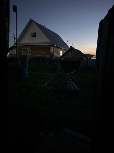 This is a low-light, outdoor photograph taken at dusk or dawn, likely in the village of Suleymanovo, Russia, given the architectural style of the wooden house and the general rural aesthetic. The scene is framed by dark wooden structures on the left and right, creating a somewhat confined perspective.

In the center of the frame is a traditional wooden house with a gabled roof. The house has light-colored siding on the upper portion and natural wood on the lower section, suggesting it is constructed from logs. A single window is visible on the upper gable, and another window with light emanating from inside is on the lower section of the house. Next to the main house, a smaller, rougher wooden structure, possibly a shed or outbuilding, is also visible. This structure has a steeply pitched roof and appears to be made of vertical wooden planks.

The foreground is dominated by dark, tall grass, with some scattered logs and a blue barrel to the left of the main house. The ground slopes upwards towards the house. In the background, power lines and utility poles are visible against a pale, fading sky. The overall impression is one of a quiet, rural setting at the edge of daylight. There are no people or animals visible in the image. The lighting suggests that artificial light sources are on inside the main house.