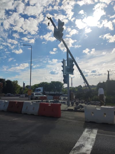 The image depicts a street construction scene in Polotsk, Belarus, under a bright, partly cloudy sky. A large crane is positioned at an angle, its boom extending over the road and lifting a traffic signal. The traffic signal is mounted on a pole, with multiple lights visible. The ground around the construction area is covered with paving stones and construction barriers, some of which are red and white. In the background, a white truck is parked, and a few individuals are visible near the construction site. A person in a white top and dark skirt is walking away from the camera in the foreground. The overall scene suggests ongoing road work or infrastructure maintenance.