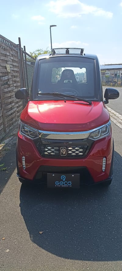 The image shows a red Geco E-AUTOMOBILE parked on a paved surface, likely a street or parking lot, in Mühlhausen, Germany. The car is facing the camera, with its front grille, headlights, and windshield clearly visible. A black license plate with the Geco logo and text "E-AUTOMOBILE" is mounted below the grille. The car has a black roof and side mirrors.
Behind the car, a wooden fence is visible on the left side of the frame, and a chain-link fence is behind that. Beyond the fences, trees and some industrial-looking buildings can be seen under a clear blue sky with scattered white clouds. A street light is positioned above and slightly behind the car. The sun is casting a shadow of the car onto the paved surface in front of it. There are no people visible in the image.