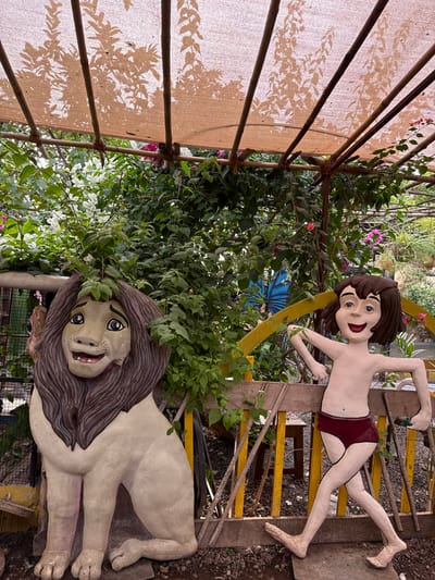 The image depicts a diorama featuring Mowgli and a lion, likely part of an amusement or garden setting, in Nashik, India. Mowgli is represented by a standing cutout figure with brown hair and red shorts. He is positioned on a wooden structure. The lion is a reclining statue, also cut out, with a tan body and brown mane. Both figures are surrounded by lush greenery, including vines and flowering plants. A partially visible awning-like structure is in the upper left, creating shadow patterns. A blue butterfly cutout is behind Mowgli. The scene suggests a tropical or subtropical climate, consistent with the location in India.