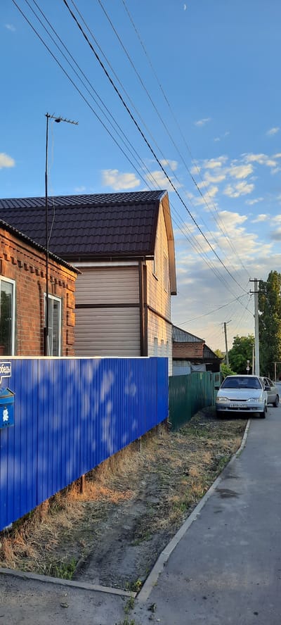 The image depicts a street scene in Shakhty City District, Russia, under a partly cloudy sky with the crescent moon visible. A residential area is shown, featuring houses with brown tiled roofs and brick or siding facades. Power lines and an antenna are prominent in the sky. In the foreground, a bright blue corrugated metal fence runs along the property line, with a blue mailbox attached to a post. Beyond the blue fence, a green corrugated metal fence is visible. Two cars are parked on the side of the asphalt road, one silver sedan in the foreground with a license plate clearly visible, and another car partially visible behind it. The ground beside the road is covered with dry grass and dirt, with tire tracks present. The overall impression is of a quiet, suburban street in the late afternoon or early evening.