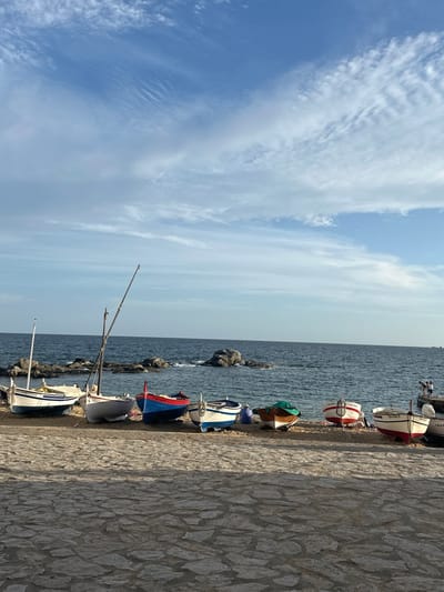 The image depicts a coastal scene in Calella De Palafrugell, Spain. In the foreground, a paved stone pathway constructed of irregular light-colored flagstones occupies the bottom quarter of the frame. Shadows fall across this pathway, originating from the right. Immediately behind the pathway is a sandy beach where eight small boats are pulled ashore, facing the sea.
From left to right, the boats are:
1. A white boat with a blue stripe and yellow trim, displaying the name "GIRONA" and registration "6-C-258-97". A mast is visible.
2. A white boat with a light blue interior, showing registration "3-CP-1-5-97", also with a mast and rigging.
3. A blue boat with a red stripe and red interior, bearing the name "PALAFRUGELL".
4. A white boat with a blue stripe, partially covered by a green tarp.
5. A dark wooden boat with a light brown interior, mostly covered by a green tarp. Two indistinct figures are visible near this boat.
6. A white boat with a red stripe, featuring a white lifebuoy attached to its bow.
7. A white boat with a red stripe and light brown interior, registered "6-CP-1-294-06".
8. A partial view of another white boat, possibly with a blue stripe, on the far right.
Beyond the beach, the dark blue sea is visible, with several prominent rock formations emerging from the water closer to the shore. Small waves break against the rocks and shoreline. On the far right, a small stone pier or jetty extends slightly into the water. Two additional indistinct figures are present on or near this pier. The sky above is clear blue with scattered, wispy white clouds, appearing denser towards the horizon. The lighting suggests either early morning or late afternoon.