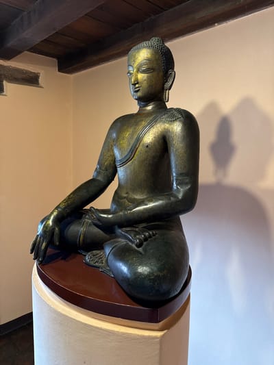A statue of a kneeling Buddha, likely made of bronze or a similar metal, is positioned indoors on a platform. The statue is of a human figure with short, curly hair, a smooth face, and clad in robes. The figure is kneeling, and one hand rests on the leg, while the other is extended. The setting is an interior space with light beige walls. A wooden beam is visible in the corner of the space. Based on the style of the statue and known Buddhist sites, it is possible this statue is located in Lalitpur, Nepal, but more information would be needed to confirm this.