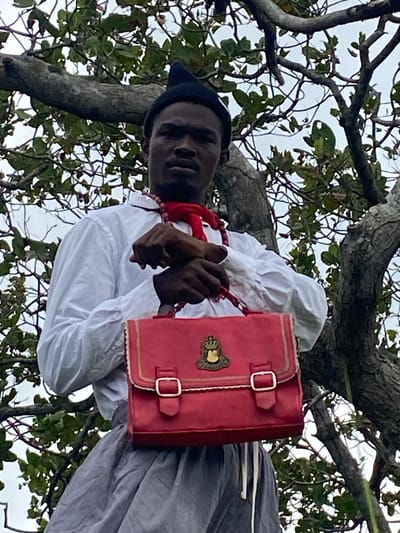 In the city of Ufu Esioni, Nigeria, a person with dark skin, wearing a black conical hat, a white collared shirt with a red scarf tied around their neck, and a grey skirt or trousers is positioned outdoors, partially obscured by the branches of a tree. The person is holding a bright red handbag with gold trim and a crest on its front. The background consists of lush green foliage and a cloudy sky.