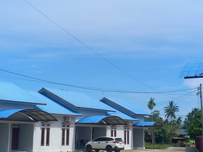 This image captures a residential street scene in Puloblang, Indonesia, under a bright, clear blue sky. The foreground features a row of modern, single-story houses, likely terraced or semi-detached units. Each house boasts a distinctive, vibrant blue corrugated metal roof and white-painted walls, contrasted with dark brown window and door frames.

Several houses are visible, with "15" and "17" clearly marked next to their entrances. Each unit has a covered carport or entrance area, some with a darker, possibly translucent, roofing material providing shade. An outdoor air conditioning unit is mounted on the wall of at least one house. In front of unit 17, a white SUV, possibly a Toyota Rush, is parked in the carport. Clothes appear to be hanging to dry near one of the houses.

The background reveals lush tropical vegetation, including prominent palm trees and other green foliage, indicative of the region's climate. Utility lines crisscross the sky, connected to a power pole on the far right. A dirt or unpaved path runs along the right side of the image, leading towards more structures and dense greenery. The bright sunlight and clear sky suggest it is daytime with pleasant weather. The overall scene depicts a calm, typical residential area.