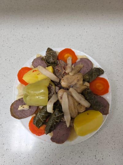 A white plate of food rests on a speckled white countertop. The plate is filled with a variety of food items including sliced carrots, what appear to be dolmades or stuffed grape leaves, pieces of cooked chicken, sliced purple sausage, boiled potatoes, and a large green bell pepper. The food is arranged in a somewhat haphazard manner. The background is a close-up of a countertop with small black speckles. The image is taken from a top-down perspective. There are no people or identifiable city locations visible in the image.