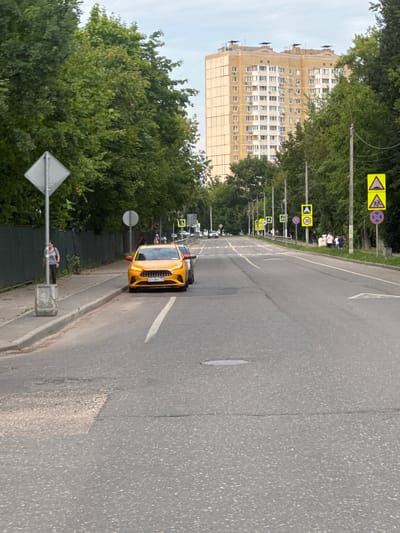 The image shows a street scene in Dedovsk, Russia. The foreground features a light yellow car parked on the right side of the road, facing away from the camera. The car has the license plate "O743MT 797". To the left of the car, a woman in a striped shirt and dark pants stands on the sidewalk, facing the street. Behind her, a diamond-shaped sign is visible. The road itself is paved and shows some wear and tear, with a manhole cover in the lower center.
The middle ground of the image displays a street lined with trees on both sides. Several traffic signs are posted along the right side of the road, including a speed limit sign (30), pedestrian crossing signs, and a general warning sign. A tall, light-colored apartment building is visible in the background, towering over the trees. A group of people are gathered on the right sidewalk, some appearing to be conversing.
The background is dominated by a cloudy sky and more trees. The overall impression is a suburban street on a cloudy day.