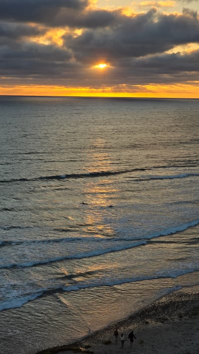 The image depicts a sunset over the Pacific Ocean at Encinitas, California. The sun, partially obscured by dark clouds, casts a golden glow across the water, illuminating the gentle waves. Two surfers are visible in the distance, paddling on their boards. Closer to shore, three figures walk along the sandy beach, their silhouettes softened by the fading light. The scene is characterized by the tranquil atmosphere of a coastal evening, with the sound of the ocean likely creating a soothing ambiance.