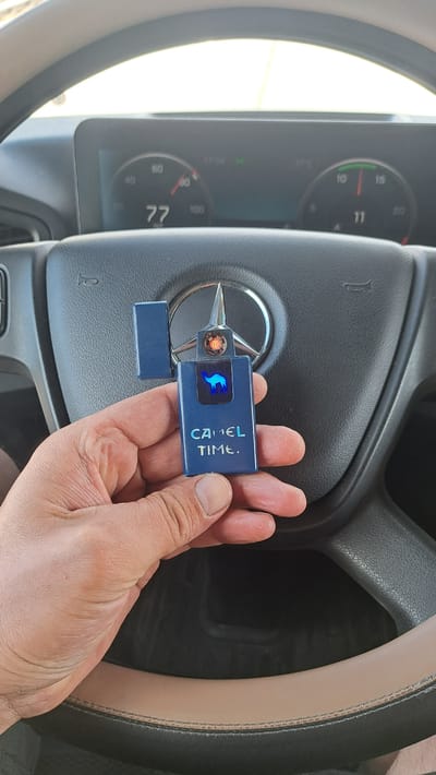 A hand holds an open, blue lighter that resembles a cigarette pack, featuring a glowing camel silhouette and the text "CAMEL TIME." The lighter is positioned in front of a black steering wheel with a silver Mercedes-Benz emblem. Visible in the background are the dashboard gauges of a vehicle, displaying a speed of 77 km/h and another gauge indicating 11. The image was taken in the city of Wenden, Germany.
