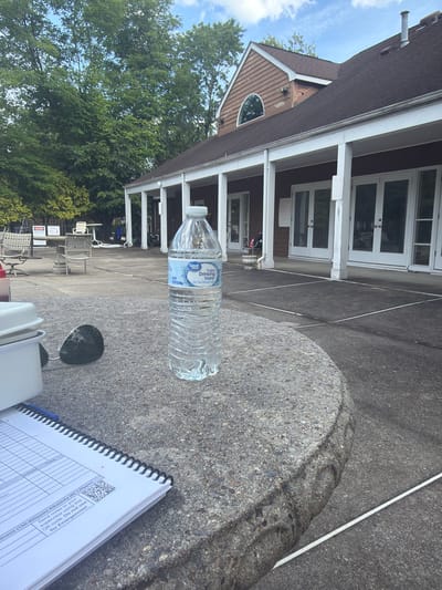 The image presents an outdoor scene in Edison, New Jersey, United States, focusing on a collection of items resting on what appears to be a stone or concrete table. A partially filled bottle of "Great Value Purified Drinking Water" is prominently placed on the table. Alongside it is a notepad, opened to a page with columns labeled "Initials" and "Time," along with text mentioning "Hardness (150-400ppm) Alkalinity (80-120ppm)" and a QR code, which is accompanied by a warning not to use it for emergencies, but rather to send notes to a supervisor. Sunglasses are also visible on the table.
The background features a building with a porch supported by white pillars and double doors, surrounded by green trees. Chairs and tables are visible on the lawn behind the table, indicating a possible outdoor leisure area. The overall setting seems to be a relaxing, outdoor environment.