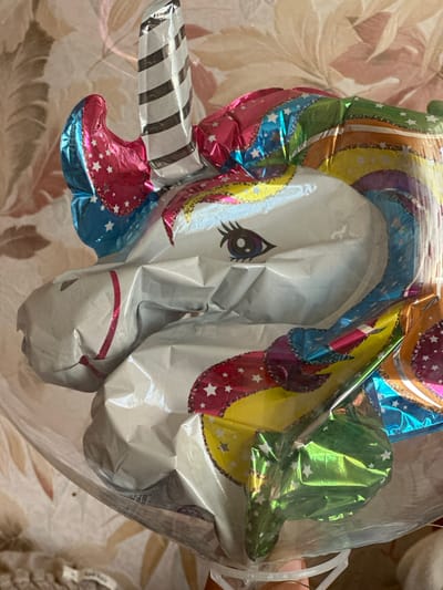 The image shows a close-up view of a white unicorn-shaped Mylar balloon. The balloon is inflated and has a colorful mane with rainbow stripes, stars, and metallic accents in pink, blue, yellow, green, and orange. The unicorn has a black eye with eyelashes, and its horn is black and white striped. The balloon is partially covered by a clear plastic bag. In the background, a floral-patterned wallpaper is visible. The bottom of the image shows a hand holding the balloon and a small piece of fabric, possibly from a stuffed animal. The image is taken in Urazovo, Russia, indicating that this is likely a personal item or decoration.
