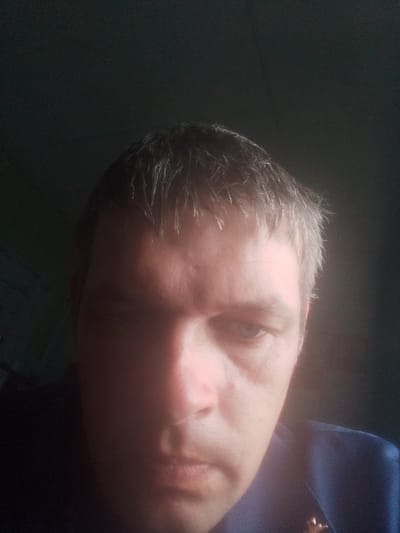 A close-up, low-angle, selfie-style photograph shows a white male in his late 30s or early 40s. He has short, light brown or graying hair, styled forward. His skin appears fair with some redness, particularly on his cheeks. His eyes are dark and appear to be looking slightly downward. His mouth is closed, with his lips pressed together. He is wearing a dark blue collared shirt or jacket, with a glint of gold-colored metal visible on his left chest, possibly a pin or emblem. The background is dark and out of focus, with a subtle texture suggesting a wall or fabric. The lighting is dim, casting shadows across the subject's face, particularly on the left side, and creating a bright highlight on his right cheekbone. The overall impression is of an indoor, dimly lit environment. This image has no discernible connection to Volchansk, Russia.