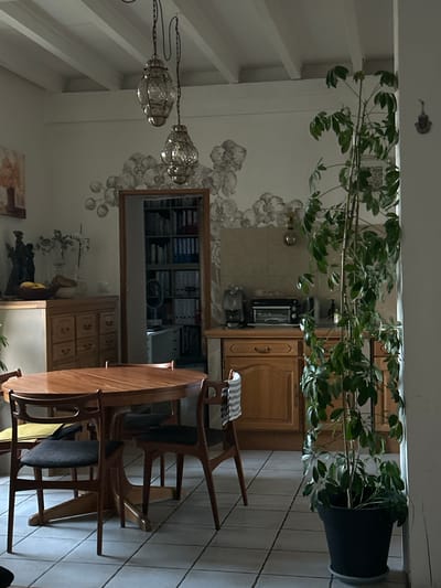 The image depicts an indoor dining area and a glimpse into a kitchen in Saint-Marcel-lès-Sauzet, France. In the foreground, a round wooden dining table with four wooden chairs with dark seats is set on a tiled floor. Behind the table, a wooden credenza is visible with several drawers and a bowl of fruit on top. To the left of the credenza, a piece of artwork hangs on the wall, and a dark sculptural figure stands on the credenza.
Further back, a doorway leads into a room filled with bookshelves, indicating a study or office area. The kitchen, to the right of the doorway, features wooden cabinets, a countertop with kitchen appliances including a coffee maker and a toaster oven.
The ceiling has exposed beams painted white. Two ornate glass and metal pendant lights hang from the ceiling. A tall potted green plant stands to the right of the scene, near the entrance to what appears to be a hallway or another room. The walls are painted white and adorned with a decorative, cloud-like mural. The overall lighting suggests it is daytime, possibly late afternoon, given the soft light. There are no people visible in the image.