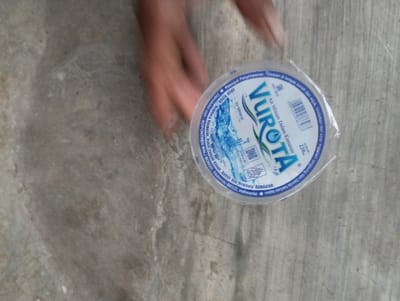 The image captures a motion-blurred hand interacting with a single-serving plastic cup of bottled water, set against a grey, textured concrete surface. The hand, visible in the upper right quadrant, appears to be either picking up or setting down the cup, indicated by the significant blur.
The plastic cup, sealed with a clear lid, prominently displays the brand name "VUROTA" in a large, blue font with a dynamic water graphic. Beneath the brand, the text "Air Minum Dalam Kemasan" (Bottled Drinking Water) is visible. Other details on the label include a barcode, the volume "220" (likely milliliters), and storage instructions in Indonesian: "Simpan di tempat bersih dan sejuk, hindarkan dari sinar matahari langsung" (Store in a clean and cool place, avoid direct sunlight). The label also specifies the product's origin, stating "produk OPPONTREN WALI SONGO, Bondowoso 68322, Indonesia," which corresponds with the provided location context of Kecamatan Panji, an area within Bondowoso, Indonesia. The overall setting is simple and functional, possibly an outdoor area or a basic indoor space, with bright, even lighting.
