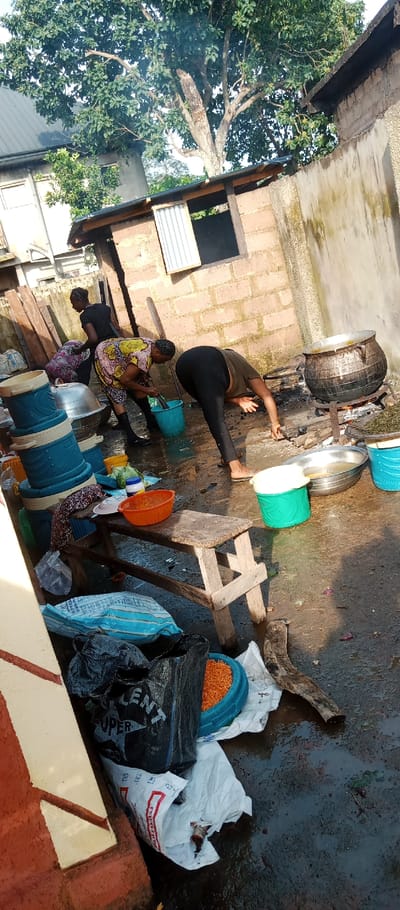 The image depicts an outdoor food preparation and cooking scene in Udi, Nigeria. Three adult females are engaged in activities on a wet, muddy, and uneven ground surface.
To the far right, a large, dark metal pot rests on a metal stand over an open wood-fueled fire, emitting smoke. It appears to contain a simmering substance. Adjacent to it on the ground is a large silver metal basin containing liquid.
In the mid-ground, one woman, dressed in a multi-colored patterned garment and dark boots, is bent over, rinsing an object in a blue plastic bucket. To her right, a second woman, in a dark top and black leggings, is bent acutely forward, attending to a silver metal basin on the ground. A smaller green plastic bucket is positioned near her bare feet.
To the far left, a third woman, dressed in dark clothing, is bent over, seemingly handling contents within a patterned fabric bag or container on the ground.
In the foreground, a rough wooden bench holds an orange plastic colander, a white plate, and green produce. Below the bench, on the wet ground, are a blue and white striped woven bag (partially labeled "SUPER TALENT"), a black plastic bag, and a blue plastic bowl containing small orange granular items. Stacked blue and cream-colored plastic coolers or food warmers are visible to the left.
The background features a building constructed of red concrete blocks with a corrugated metal roof and a small window frame, partially obscured by a large tree with dense green foliage. To the right of this structure is a stained concrete wall.