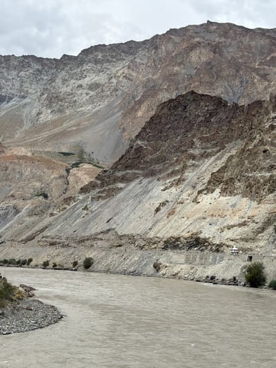 The image depicts a wide, muddy river flowing through a valley with steep, rocky mountains on either side. The mountains are arid and appear to be composed of layers of sedimentary rock. The riverbank on the left is lined with small pebbles and some sparse vegetation. On the right bank, there is a road carved into the mountainside, with a small white structure, possibly a stupa or shrine, and a small building next to it. The sky is overcast with clouds. The scene is likely located in a high-altitude region, given the rugged terrain. The location is identified as Chilling, India.