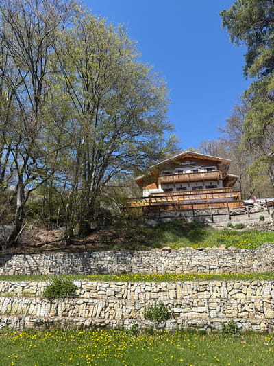 The image shows a building in Ichera, Bulgaria with a stone wall running along the side of it. The building is three stories tall and has a wooden exterior. There is a person standing on the balcony of the top floor. A large tree is in the foreground. The sky is blue.