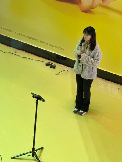 This image captures a scene within an indoor commercial or event space, located in Changping District, China. The environment is characterized by uniformly bright yellow walls and floor. A prominent yellow wall displays Chinese text advertising cosmetic products, specifically "Willow Silk Wood Moisturizing Light & Transparent Sunscreen" and "Willow Silk Wood Vitality Light-Blocking Whitening Sunscreen", alongside a partial large graphic of what appears to be a human arm or hand in the upper section. A single female individual stands centered in the frame. She has dark, shoulder-length hair and wears glasses. Her attire consists of a light grey textured jacket worn over a lighter garment, dark trousers, and dark, light-accented shoes. Her hands are engaged in front of her midsection, appearing to adjust something. To her left, a black tripod stand supports a tablet holder angled upwards. On the yellow floor, behind the tripod, two small black electronic devices with attached cables are visible, indicating audio or broadcasting equipment. The overall arrangement suggests a setting for a public address, recording, or live broadcast.