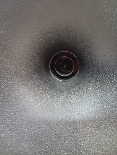 The image shows a close-up, overhead view of a drain in a textured, gray surface. The drain itself is circular with concentric rings. The surface appears to be some kind of metallic or plastic material with a rough, dimpled texture, possibly a sink or a tub. The lighting is from above, casting a slight shadow into the drain. The location is not specified in the image or crops.