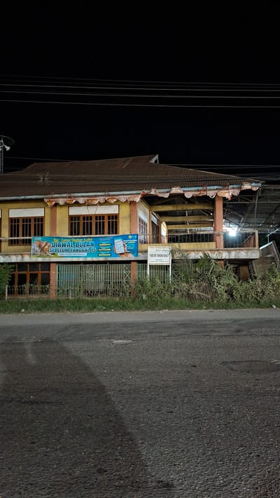 The media file shows a building with a yellow facade and a brown corrugated metal roof, likely at night given the dark sky. The building appears to be a commercial establishment or a residential building with multiple windows. There are several vertical lines that could be power lines or structural elements, against the dark background.
In front of the building, there is a sign in Indonesian that appears to be an advertisement or announcement. It's a vertical banner with blue and orange elements and text. The text visible on the banner includes "DIAWAL BULAN", "SEBELUM TANGGAL 15", and "LEBIH TERANG, LEBIH NYAMAN". There is also a smaller white sign on a post to the right of the banner with Indonesian text, possibly indicating the name of the establishment or location, reading "PADIS SU TAMPING BANGSA".
A paved road runs alongside the building, with a grassy area and some vegetation between the road and the building. The overall scene suggests an urban or semi-urban setting in Keudeteupinpunti, Indonesia. The lighting from within the building and a street lamp illuminates parts of the facade and the road. There are no people visible in the media file. The weather appears to be clear, with no indication of rain or extreme conditions. The time of day is clearly evening or night, judging by the dark sky and artificial lighting.