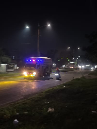 An exterior, eye-level, night-time, medium shot captures a road in Tanahterban, Indonesia, illuminated by streetlights and vehicle lights. In the foreground, a grassy embankment lines the right side of the road, with some litter visible. The road itself is wet and reflects the lights. A yellow truck with blue and pink lights on its roof is the primary subject, moving from left to right across the frame. Its headlights are on, casting a bright yellow glow. To the right of the truck, a motorcycle rider, illuminated by the truck's lights and their own headlight, is also moving from left to right. In the background, more streetlights are visible, along with other vehicles and buildings. The scene suggests activity on a road at night.