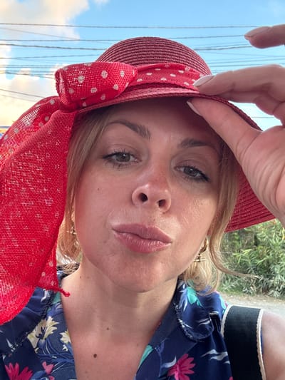 This is a close-up, rotated portrait of a woman wearing a red, polka-dotted hat with a wide brim and a bow. She has blonde hair and is looking towards the camera. Her skin has a light tan with some freckles. She is wearing a blue dress with a floral pattern of pink and teal flowers. Her hand is visible, holding the brim of her hat. The background shows a bright sky with some thin, dark lines that could be power lines, and out-of-focus green foliage. The lighting suggests it is daytime. The overall impression is casual and outdoorsy.