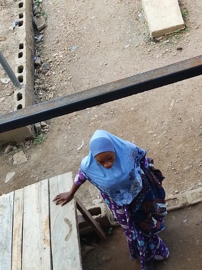 In Abeere, Nigeria, a woman wearing a light blue hijab and a patterned blue and purple dress stands near a wooden surface. The woman's hand is placed on the wooden surface, and she is looking downwards. The setting appears to be outdoors, with dirt and gravel on the ground. There are also some construction materials visible, including cinder blocks and a metal beam. The scene suggests a casual or everyday moment, possibly related to an outdoor market or a work area. There is no visible text in the image, and the time of day and weather conditions are not explicitly indicated, though the lighting suggests it is daytime.