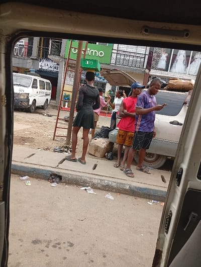 The image provides a street-level view of a bustling urban scene in Rumuokoro, Nigeria, captured from within a vehicle, with its frame visible on the top, left, and right. The setting appears to be a commercial area or market, characterized by a mix of concrete and dirt surfaces, and scattered litter on the sidewalk and road.
In the mid-ground, a person, possibly a young man, stands facing away from the camera, wearing a dark, short-sleeved dress with sheer long sleeves and slippers. They are carrying a green plastic crate balanced on their head. Nearby, a tall, red-painted metal ladder stands upright, next to a small display of blue artificial flowers.
To the right, a group of men are gathered. One wears a red t-shirt and brown shorts, while another in a purple and black striped t-shirt and black shorts is preoccupied with a smartphone. A third man in a pink shirt is partially visible behind them. All wear sandals or slippers. Behind this group, the trunk of a light-colored car is open.
In the background, a white van, resembling a minibus, is parked on the left, displaying the number "2114" and partially legible text that appears to be "PACIFIC GOODS". Buildings line the street, including one with a prominent green sign showing "Otim...mo", likely "Optimo". Another building on the far right showcases wedding dresses in its windows.
The scene depicts everyday activity under natural, diffuse daylight, suggesting either overcast weather or mid-morning/afternoon. The people are casually dressed, indicating a relaxed atmosphere amidst the urban bustle.