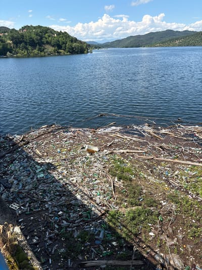This media file captures a stark contrast between natural beauty and severe environmental pollution at Muhovo, Bulgaria. The image, presented with a vertical orientation, depicts a wide body of water, likely a river or lake, with undulating ripples reflecting the clear blue sky. On the opposite side of the water, lush green, tree-covered hills rise under a bright sky dotted with fluffy white clouds, suggesting a serene natural landscape.

However, the foreground reveals a heavily polluted shoreline. The bank is almost entirely covered with an dense accumulation of trash, predominantly plastic bottles of various sizes and colors, along with glass bottles, plastic bags, styrofoam fragments, and natural debris like branches and twigs. Patches of green grass and small plants attempt to grow amidst the garbage, highlighting the extent to which the natural environment has been overwhelmed by human waste. The water's edge, where it meets this refuse-strewn bank, is also choked with floating debris.

The scene takes place during daylight hours, under a sunny sky, as indicated by the clear lighting and shadows cast on the ground. There are no people visible, and no specific activity is taking place other than the static display of accumulated pollution. The image serves as a powerful visual testament to environmental degradation, with the vast quantity of discarded items hinting at a systemic issue of improper waste disposal. No distinct, legible text is visible on any of the discarded items.
