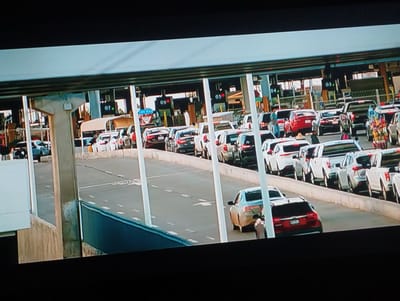 The image shows a line of cars waiting at a toll booth. There are several lanes, each marked with a number (01-07). Some people are standing around the cars. The toll booth area is covered by a large roof structure supported by white columns. The scene appears to be during the daytime. This is not likely in the city of Punta de Piedras, Venezuela.