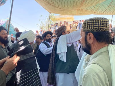 A group of men are gathered under a canopy at a location in Zaker-E Sharif, Afghanistan. The men are wearing traditional clothing, including long white shirts, turbans, and black skullcaps. The men are interacting with each other, and one man is seen adjusting the turban of another man in the foreground. The scene is likely taking place at a public gathering or ceremony. The men are in the foreground of the image, while the background is filled with a large number of people who are also wearing traditional clothing. The scene is located in Zaker-E Sharif, Afghanistan, which is known for its large religious community and many mosques.