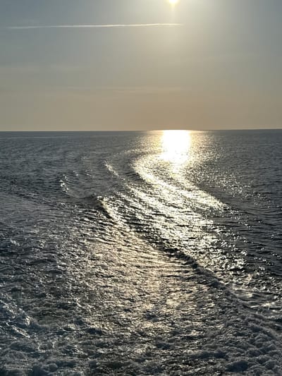 The image depicts an expansive view of open sea and sky, observed from the waters adjacent to Palaiopoli, Greece. The sun is prominently situated in the upper-right sky, casting a brilliant, elongated reflection that traverses the water surface from the distant horizon to the immediate foreground. The sky exhibits a gradient from a light blue in the upper portion to a warm, pale yellow-orange hue near the horizon line. A singular, thin, horizontal contrail or cloud is discernible in the upper-left sky. The ocean surface, colored dark blue-grey, features a distinct, curvilinear wake of agitated, foamy water originating in the lower-left and extending towards the center-right, indicative of the rearward propulsion of a moving vessel. The wake dissipates into a series of ripples further from the foreground. No land formations, additional vessels, or human subjects are present in the visible frame.