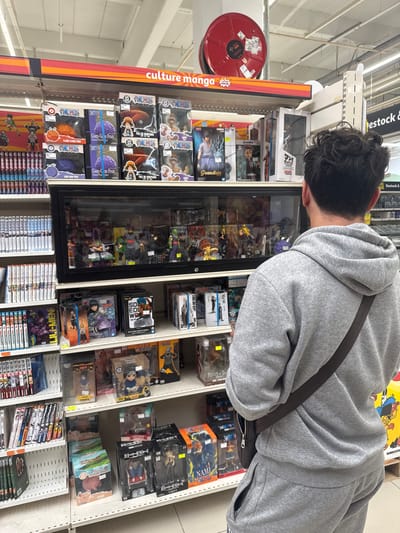 The image captures an indoor retail environment, specifically a store selling manga and anime merchandise, located in Leers, France. In the foreground, a young male with dark curly hair, wearing a grey hooded sweatshirt and sweatpants, is viewed from behind, looking at a display shelf. His right hand is holding a dark brown purse or messenger bag strap.
The shelves are filled with various collectible items, primarily action figures and manga volumes. Prominently displayed on a shelf above the main display case are "One Piece" themed items, including figures of characters and "Devil Fruit" replicas. The main display case contains a collection of action figures, some of which appear to be from popular anime series such as "Naruto" and "Dragon Ball." Below this, more shelves are packed with boxed figures and manga books.
The store environment is well-lit, with fluorescent lighting on the ceiling. A red fire hose reel is visible mounted on the wall above the "culture manga" signage. Another sign on the right side of the image reads "stock &". The overall scene suggests a retail setting focused on pop culture collectibles.