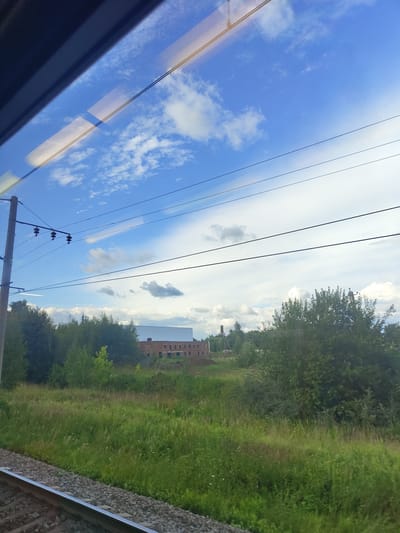 The image shows a view from a train window. In the foreground, there are train tracks. The middle ground is a grassy field with trees. In the background, there is a building under construction with a white roof and brick walls. There are also power lines stretching across the sky. The sky is blue with white clouds. The image appears to have been taken in Фруктовая, Russia.