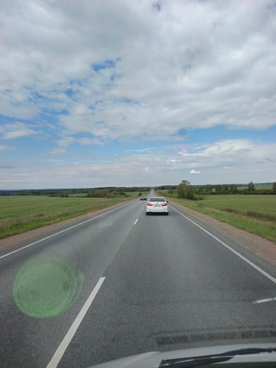 A white sedan with the license plate E825HT154 is traveling on a two-lane asphalt road, heading away from the viewer. The road is bordered by green fields and trees. The sky is partly cloudy with a mix of blue sky and white clouds. The image appears to be taken from another vehicle, with a portion of the windshield visible in the foreground. The scene is likely from the city of Kovriga, Russia.