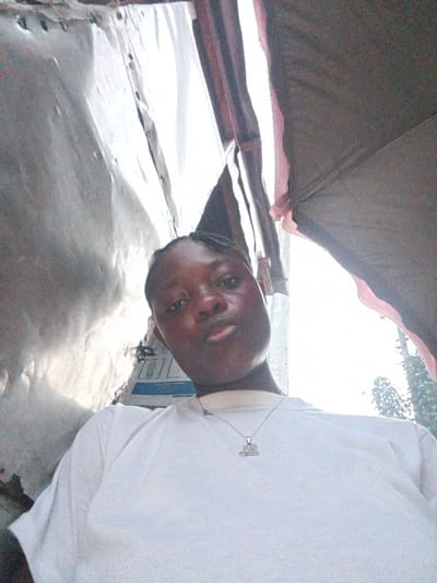 A low-angle self-portrait features a young woman with dark skin and short, braided hair, looking directly at the camera with a neutral expression and slightly puckered lips. She is wearing a plain white short-sleeved t-shirt and a silver necklace with a rectangular, block-letter pendant, the specific word or design on which is not clearly discernible.
The background suggests an informal, possibly makeshift indoor or semi-enclosed setting. To the left of her head, a section of corrugated metal sheeting is visible, reflecting ambient light. To the right, a darker, possibly fabric or tarp-like material forms part of the ceiling or upper wall. A very bright light source, likely natural sunlight, is prominent behind her head and to her right, illuminating what appears to be an opening to the outside, with a faint suggestion of green foliage.
The lighting highlights parts of her face, such as her forehead and cheeks. The overall impression is an informal snapshot taken during the daytime in a simple environment, consistent with a location such as Mbribit, Nigeria.
