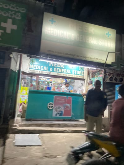 The image shows a street-level view of a storefront at night, identified as "MEER MEDICAL & GENERAL STORE" in B Camp, India. The storefront features a large, illuminated sign with the store's name and a plus symbol, indicating a pharmacy or medical supply store. The entrance to the store is open, revealing shelves stocked with various products, and at least one employee is visible behind the counter. A teal-colored counter or display unit is positioned in front of the store. Several individuals are present in the foreground and on the street, some appearing to be customers or passersby. A motorcycle is parked in the lower right portion of the frame. The overall scene suggests a busy commercial area, with the lighting indicating it's after sunset.