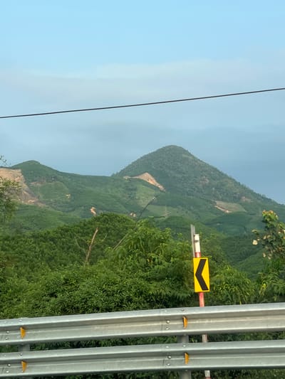 A vertical outdoor shot in Aê Thi, Vietnam shows a view from a hillside road with a metal guardrail on the right. A yellow and black chevron sign is attached to the guardrail, indicating a sharp curve. Lush green vegetation covers the hillside, extending to the horizon. In the distance, a mountain peak can be seen under a hazy sky. A power line is visible on the left side of the frame, cutting diagonally across the sky.