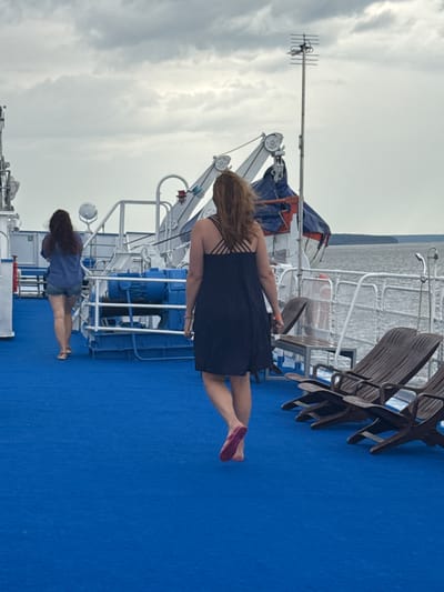 The image depicts a blue deck on a ferry, likely in or near Belous, Russia, with a cloudy sky overhead. In the foreground, two women are walking away from the camera. The woman on the right wears a dark blue dress and pink flip-flops. The woman on the left wears a blue shirt and denim shorts. Behind them, lounge chairs are arranged on the deck. Further in the background, the railing of the ferry and the water are visible. The scene appears to be a calm, overcast day on a ferry.