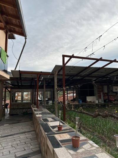 The image shows an exterior scene, possibly a backyard or patio in Petrovo, Bulgaria. A metal structure, seemingly a support frame for solar panels, dominates the view. The sky is overcast. A vine is strung along wires near the metal frame. In the background, a brick wall and some electrical boxes are visible. The patio area is paved with patterned stones, and a ledge holds several small potted plants with metal stems. In the lower left corner, a section of a building roof is visible.